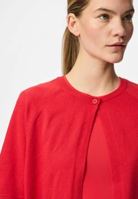 Crvena cardigan jakna s okruglim izrezom, zatvaranjem na dugmad i raglan rukavima. Mekana tekstura; lagana tkanina; složena preko odgovarajuće majice.