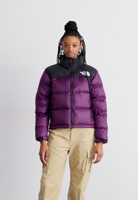 Puffjakke med en lilla kropp, svarte skuldre, glidelås og elastiske mansjetter; kombinert med beige cargo-bukser.