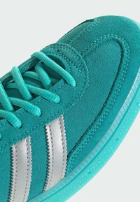 Baskets en daim bleu sarcelle avec lacets turquoise, accents à rayures argentées et semelle en caoutchouc turquoise texturée avec un motif géométrique.