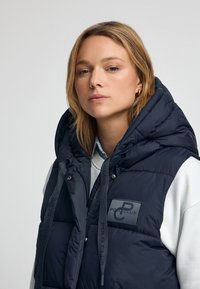 Marinblå puffer-väst med en stor huva, med framsidans dragkedja, logotyp-patch och grå dragsnören, bärs över en vit sweatshirt.