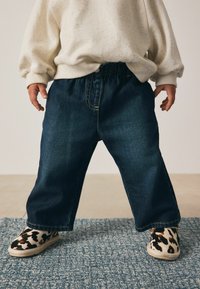 Jeans v temno modri barvi z elastičnim pasom; oblečeni z lahkim kremnim puloverjem in črnobrnimi čevlji s potiskom.