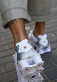 Ténis brancos com detalhes prateados, parte superior em tecido, sola robusta, marca visível e detalhes vermelhos na sola exterior. Tecido texturizado perto do calcanhar.