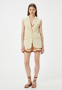 Beige ærmeløs blazer med to frontlommer og knaplukning, parret med brune tie-dye shorts og brune snørebåndsandaler.