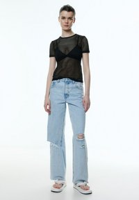 Schwarzes Mesh-T-Shirt mit kurzen Ärmeln kombiniert mit hellblauen, ausgewaschenen Schlagjeans; Model trägt weiße Flip-Flops und minimale Accessoires.