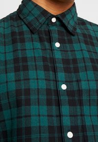 Grün-schwarz kariertes Hemd mit Button-Down-Kragen, weißen Knöpfen, weicher Textur und sichtbaren Nähdetails.