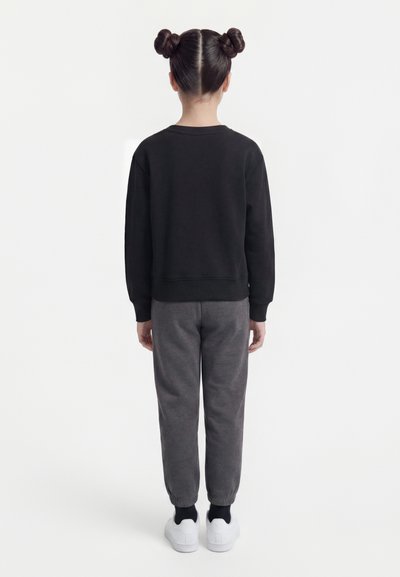Παιδί με δύο κότσους στα μαλλιά που φοράει μαύρη μπλούζα με μακριά μανίκια, γκρι παντελόνι φόρμας, μαύρες κάλτσες και λευκά αθλητικά παπούτσια, στέκεται γυρισμένο με την πλάτη.