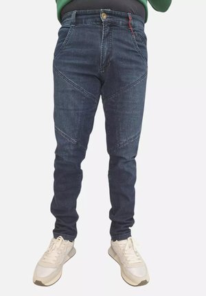 Jeans slim fit blu scuro con dettagli diagonali cuciti, indossati con sneakers bianche da una persona in piedi contro uno sfondo bianco.