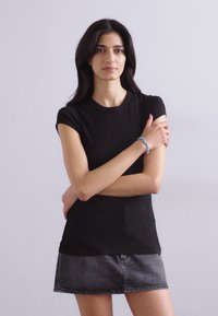 Čierne priliehavé tričko s krátkym rukávom z hladkej látky, doplnené šedou denimovou minisukňou. Modelka má prekrížené ruky a nosí modrý náramok.