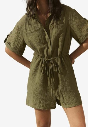 Mujer con mono corto texturizado de manga corta en verde oliva, con botones en la parte delantera y cintura ajustable con cordón, de pie con las manos en las caderas.