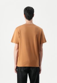 T-shirt en coton marron avec col rond, manches courtes et coupe décontractée, associé à un jean en denim noir. Coutures et étiquettes minimales visibles.