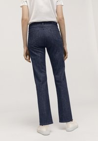 Jeans acampanados de denim azul oscuro, con una cinturilla estándar, dos bolsillos traseros y costuras visibles, combinados con zapatos blancos.