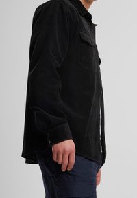 Chemise en velours côtelé noir avec fermeture boutonnée sur le devant, deux poches poitrine, manches longues et ourlet légèrement arrondi. Le tissu a une finition texturée.