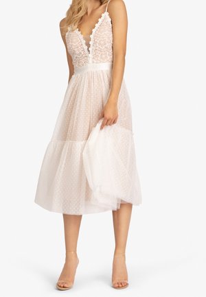 Femme portant une robe blanche mi-longue avec un corsage en dentelle florale, un décolleté en V profond, des bretelles fines et une jupe transparente à pois, tenant une partie du tissu.
