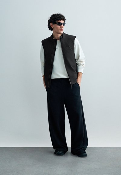 Fekete ujjatlan dzseki világosszürke pulóver felett, széles fekete nadrággal párosítva. A modell sötét napszemüveget visel, világos háttér előtt áll.