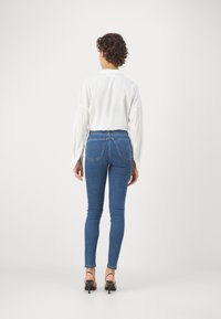 Jeans ajustados de mezclilla de color azul, con dos bolsillos traseros, combinados con una camiseta de manga larga blanca y suéter suelto, y zapatos de tacón negro.