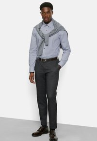 Hemd mit blauem geometrischen Muster, grauer Pullover über die Schultern gebunden, graue Anzughose und braune Lederschuhe.