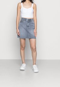 Woman wearing white cropped tank top, gray denim mini skirt with frayed hem, and white sneakers, standing against plain background.