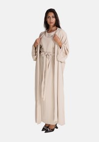 Beige kimono-achtige badjas met lange mouwen en een striktaille, gelaagd over een bijpassende jurk. Glanzende zwarte puntige hakken met strikaccenten.