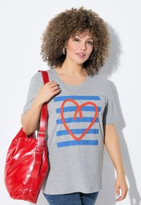 Camiseta de algodón gris con rayas azules y un diseño de corazón rojo, combinada con un bolso tote rojo brillante. Modelo con cabello rizado y aretes.