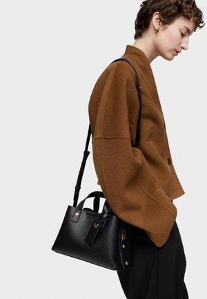 Mujer con una chaqueta marrón de gran tamaño y pantalones negros, llevando un bolso de mano de cuero negro con una correa para el hombro en su lado izquierdo.