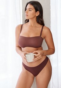 Braunes Bikini-Set mit einem Bandeau-Oberteil mit schmalen Trägern und hochgeschnittenen Bottoms, gefertigt aus glattem, dehnbarem Material. Hält eine Keramiktasse.
