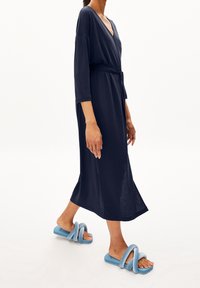 Granatowa, średniej długości sukienka kopertowa z dekoltem w kształcie litery V i długimi rękawami, zwężająca się w talii za pomocą wiązania. W zestawie z jasnoniebieskimi, pluszowymi klapkami.