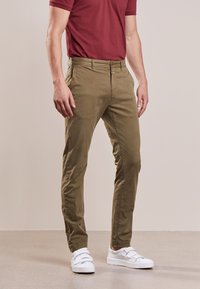 Chinos vert olive avec une texture lisse, coupe slim et fermeture éclair. Associés à des baskets blanches, créant un look décontracté.