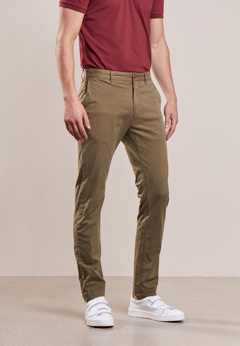Chinos vert olive avec une texture lisse, coupe slim et fermeture éclair. Associés à des baskets blanches, créant un look décontracté.