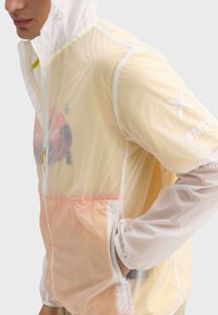 Veste à capuche légère en nylon transparent. Présente un tissu dégradé de l'orange au jaune, une fermeture éclair et des accents imprimés sur les manches.