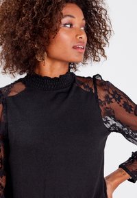 Pull noir avec un col smocké haut. Les longues manches sont en dentelle noire transparente ornée de motifs floraux. Texture de tissu douce.