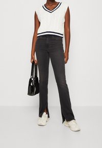 Bílé bezrukávové svetříkové tričko s úpletem a námořnickými pruhy na límci, v kombinaci s černými vyššími zvonovými džíny se bočními rozparky. Drží černou kabelku.