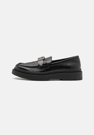 Mocassin en cuir noir avec semelle épaisse, boucle argentée décorative et détail cousu sur le devant, incliné vers la gauche sur fond uni.