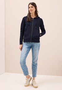 Sweatshirt zippé marine avec deux poches avant, capuche à cordon de serrage et poignets à côtes. Porté avec un jean bleu clair et des baskets beiges avec des accents bleus.