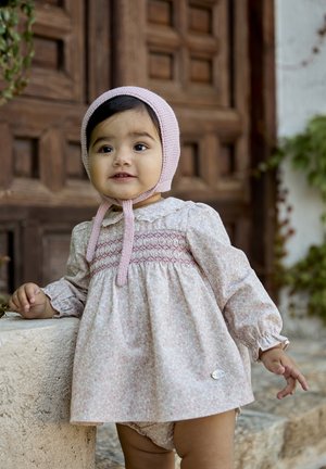 Vestido floral de color rosa claro con fruncido, mangas largas abullonadas y un cuello blanco; combinado con una bonete de punto rosa, fondo de piedra texturizada.