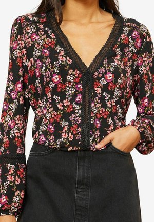 Femme portant un chemisier noir à fleurs roses, rouges et blanches, bordure en dentelle au col en V et aux manches, rentré dans une jupe noire. Une main dans la poche.