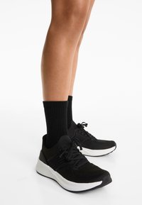 Zwarte sportschoenen met een gemêleerde bovenkant van gebreid textiel, witte tussenzool en zwarte veters, gedragen met zwarte geribbelde sokken.