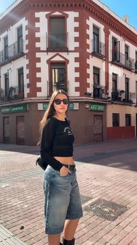 Νέα γυναίκα που φοράει γυαλιά ηλίου, μαύρο crop top και τζιν σορτς στέκεται σε ηλιόλουγη γωνία δρόμου με κτίριο από τούβλα και μπαλκόνια πίσω της.