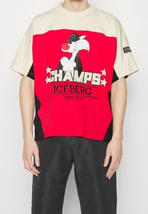 Personne portant un t-shirt rouge et beige avec un chat cartoon, le texte «CHAMPS ICEBERG depuis 1974» et un écusson noir sur la manche.