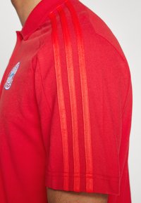 Rød polo-shirt med korte ærmer, der har tre mørkerøde lodrette striber på skulderen og et cirkulært logo på brystet.