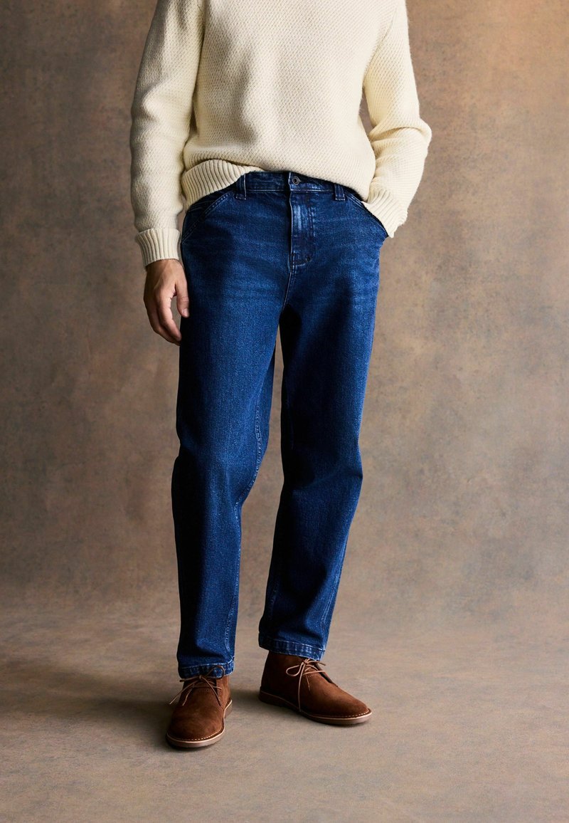 Jeans de mezclilla azul oscuro con un corte relajado, combinados con un suéter de punto color crema y zapatos de ante marrón. El fondo es neutro y texturizado.