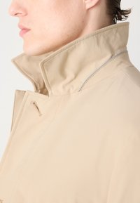 Beige színű, könnyű dzseki magas gallérral, sima felülettel és diszkrét szürke varrással a gallér szélén.
