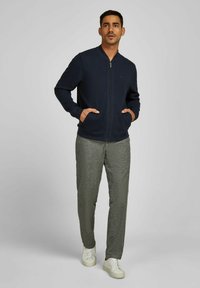 Marinefarbener Reißverschluss-Pullover mit langen Ärmeln und seitlichen Taschen, kombiniert mit strukturierten grauen Hose und weißen Sneakern. Schlichtes, modernes Design.