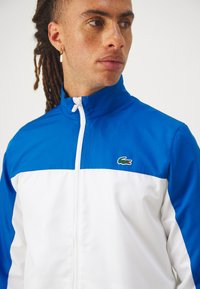 Zila un baltā krāsā ražota jaka ar rāvējslēdzēju, ar augstu apkakli, gludu audumu un nelielu Lacoste krokodila logotipu kreisajā krūtī.