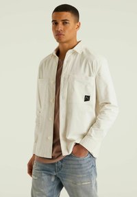 Weißes Hemd mit Knopfleiste und zwei Brusttaschen, mit einem kleinen schwarzen Logo. Trägt man über einem hellbraunen T-Shirt und kombiniert mit blauen, distressed Jeans.