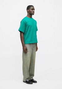 Πράσινο κοντομάνικο μπλουζάκι με τσέπη στο στήθος, συνδυασμένο με φαρδιά, ανοιχτόχρωμα πράσινα τζιν και μαύρα παπούτσια, τοποθετημένο όρθιο σε απλό φόντο.