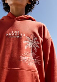 Rust-colored hoodie with front pocket; features white embroidered text "LOS ANGELES CALIFORNIA" and a palm tree with sun and surfboard design.