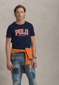 Tengerész kék póló "POLO" logóval, koptatott kék farmernadrággal, amelyen foltok találhatók, és egy narancs színű kötött pulóver a derekán megkötve.