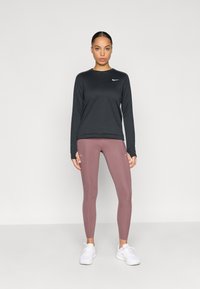 Haut de sport noir à manches longues avec un col rond et un logo blanc ; associé à des leggings moulants mauve. Des chaussures de sport blanches complètent le look.
