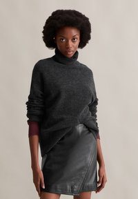 Dunkelgrauer Rollkragenpullover, gestrickte Textur, lockerer Schnitt, kombiniert mit einem schwarzen Lederwickelrock, der eine glatte Oberfläche und minimale Nähte zeigt.