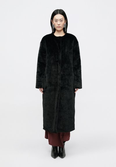 Schwarzer Kunstpelzmantel, lang und übergroß, mit rundem Ausschnitt und ohne Verschlüsse. Das Modell trägt einen maroonfarbenen Rock und schwarze Stiefel mit Absatz.